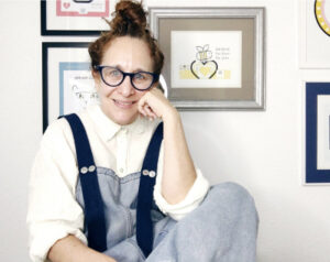 Artist photo of Sharon Glassman seated on chair smilng in front of wall of Smile Songs art prints