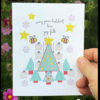 hand holding holiday card with trees, bees, stars and gifts in front of field of flowers