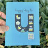 happy u day card held by hand against greenery to show art and qr code that plays ong